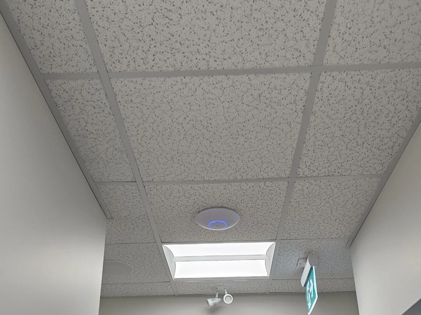 A ceiling-mounted Ubiquiti Wi-Fi access point installed by KeenAI in a commercial ceiling grid alongside an LED troffer and an emergency exit light