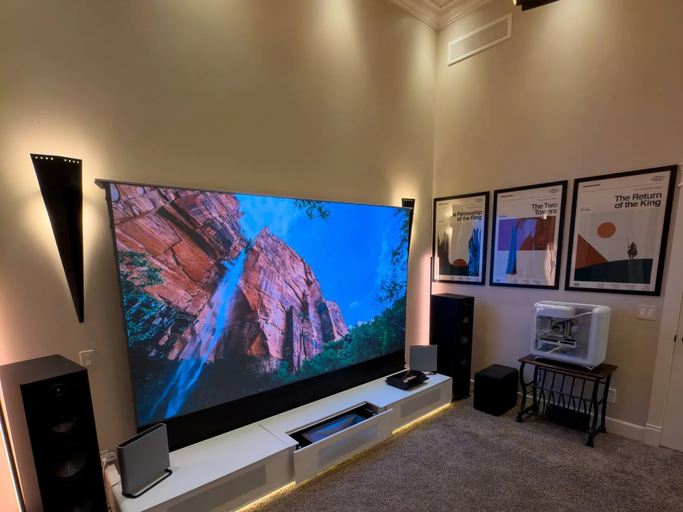 A home theatre installed by KeenAI: a large drop-down projection screen flanked by floor-standing speakers and wall sconces, with concealed cove lighting along a low white credenza