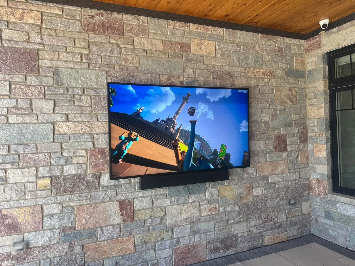 An outdoor-rated television and soundbar mounted on a stone wall under a covered patio by KeenAI, with a dome security camera on the same soffit