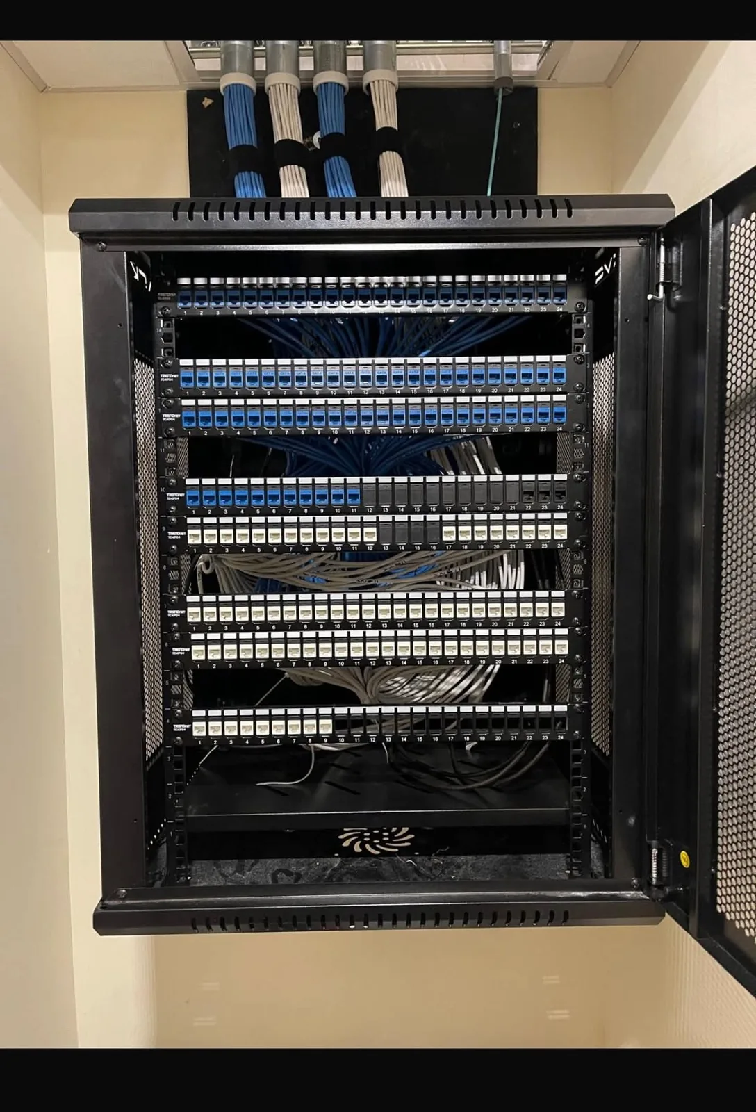 A wall-mounted equipment rack installed by KeenAI with eight patch panels of numbered Cat6 terminations and blue and white cable bundles dressed through ceiling conduit